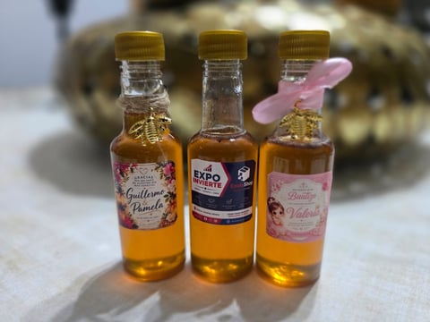 Three small glass bottles with golden caps containing amber liquid, arranged with a pink spoon, and a blurred succulent plant in the background