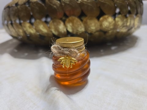 Small glass jar of golden honey with a gold metal bee charm and twine bow, decorative gold textured vessel in background