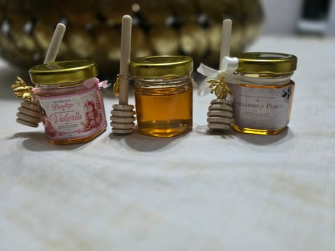 Three small glass jars with honey dippers and decorative wooden beads, containing different colored liquids and honey varieties