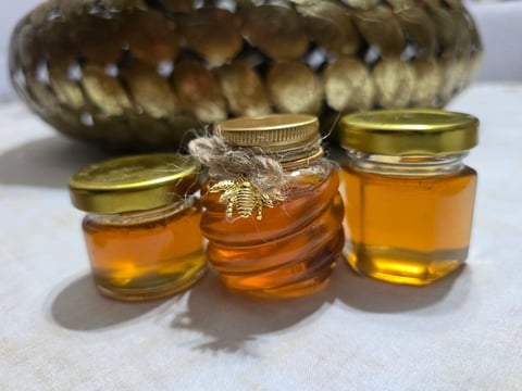Three small glass jars of honey with gold lids and twine bows, arranged in front of a woven basket on a white surface
